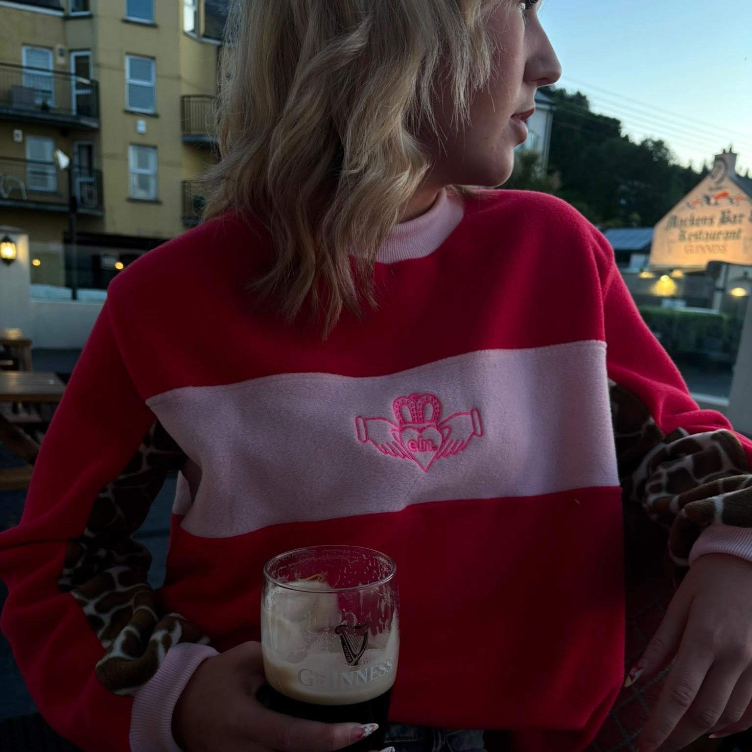 Person wearing a red and white sweater with a logo, holding a drink outdoors.