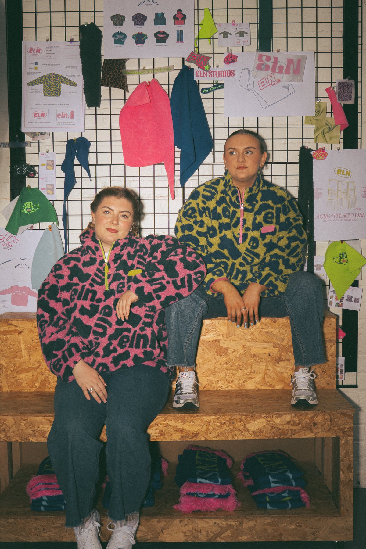 Two women wearing matching pink and green print half zip fleeces sit on a wooden bench in front of a grid wall covered in fashion sketches and pinned garments.